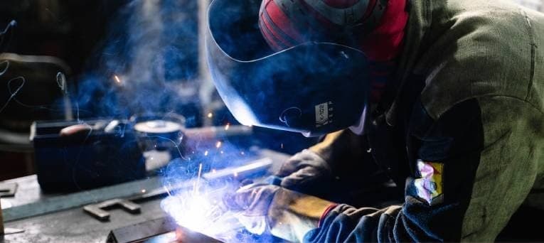 A welder in a heavy jacket burning a rod at a bench, blue smoke rising
