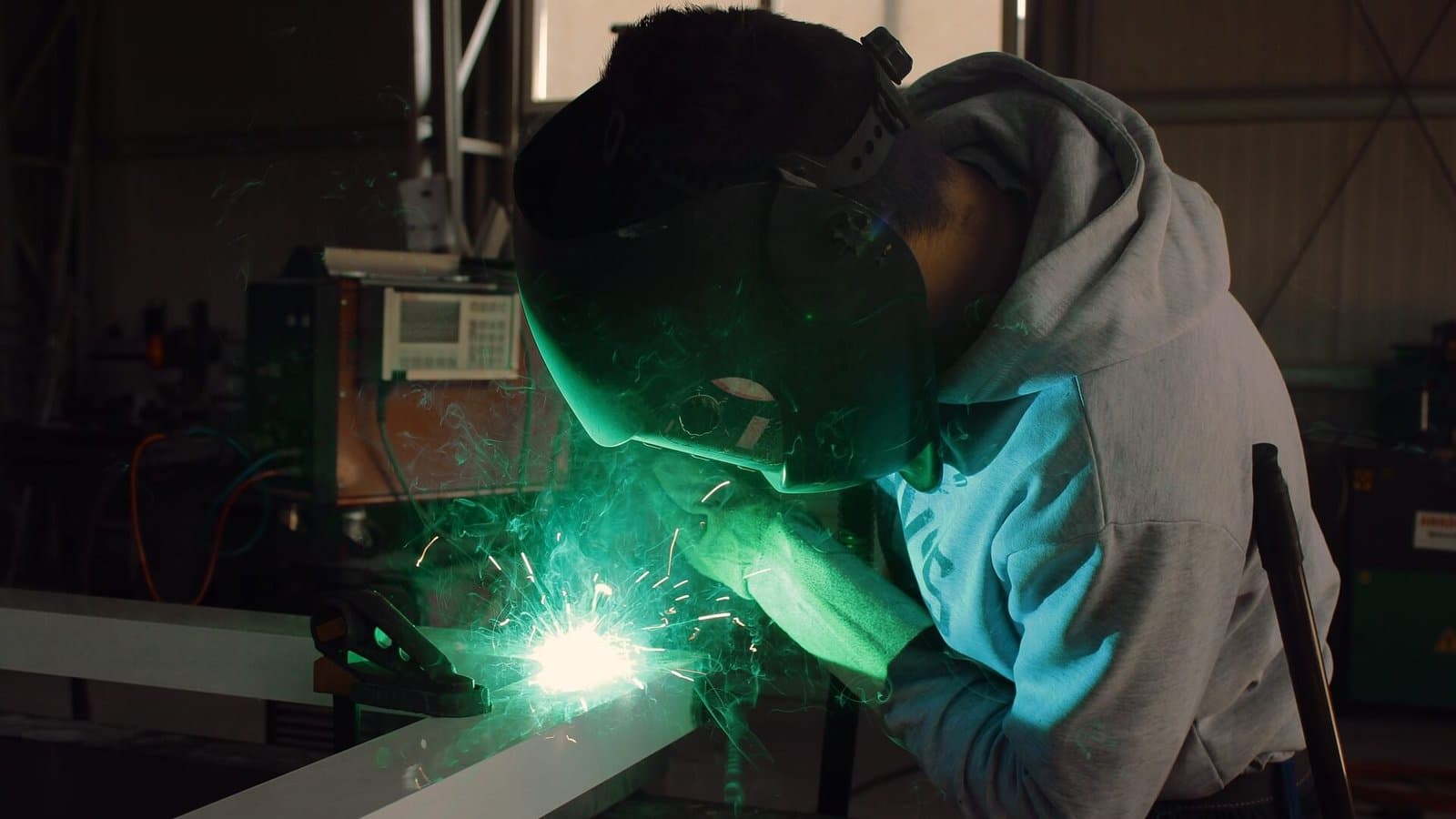A welder leaning over a steel beam, the arc lighting the workshop green