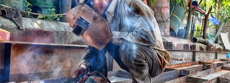 Two welders crouched over a steel beam on an outdoor site, smoke rising from the joint