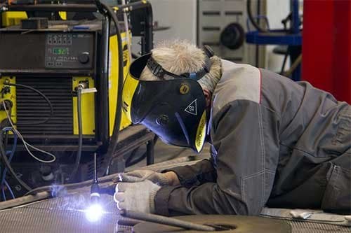 A welder in a helmet laying a bead on steel tubing at a workshop bench