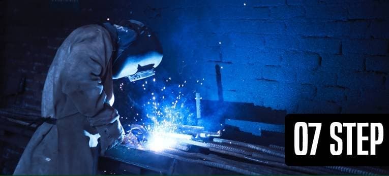 A welder in a canvas jacket working over a bench of steel bar, sparks flying against a blue-lit brick wall