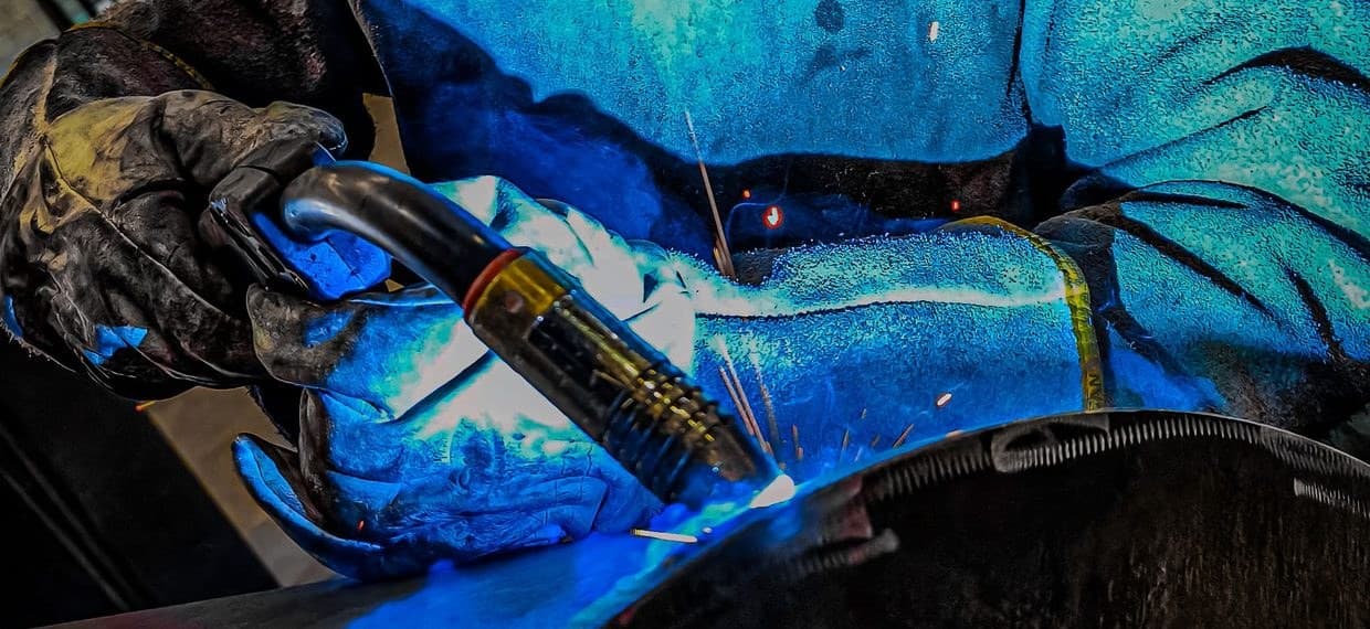 A gloved hand running a wire-feed welding gun along a steel seam, sparks scattering across the welder's leather sleeve
