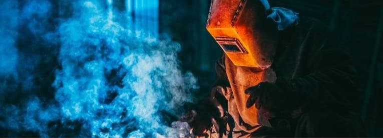 A welder in a hood working through a cloud of blue welding fume