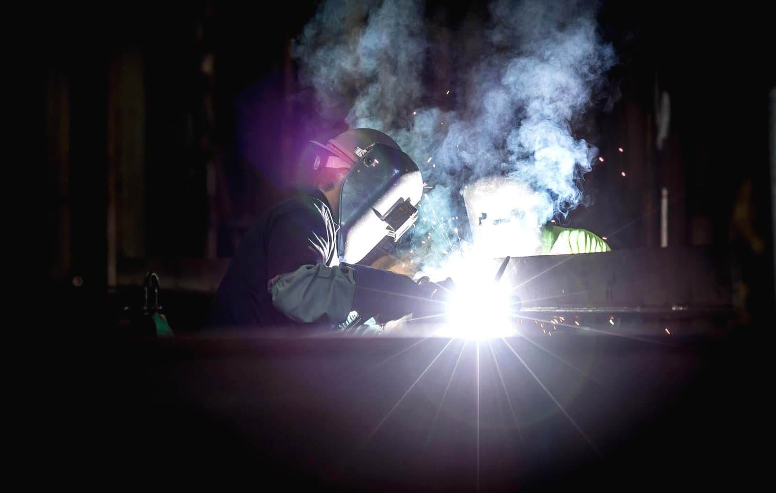 A welder at a bench in a dark shop, the arc flaring bright white