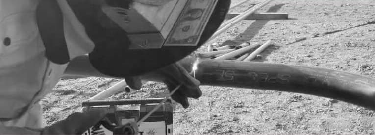 A black and white close-up of a welder joining steel tube on the ground