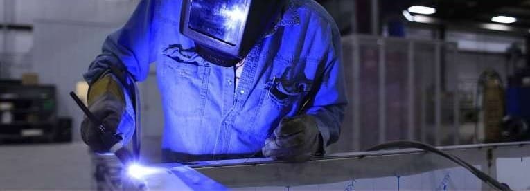A welder in a blue jacket running a bead along a steel bar at a bench