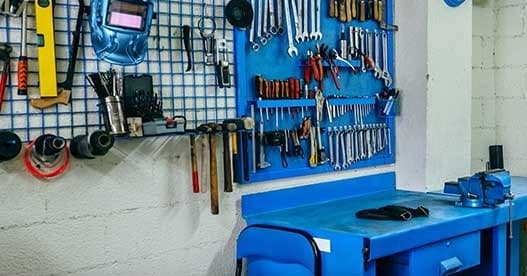 A blue welding helmet and a row of hammers hanging on a workshop pegboard, above a steel bench with a bolted-down vice