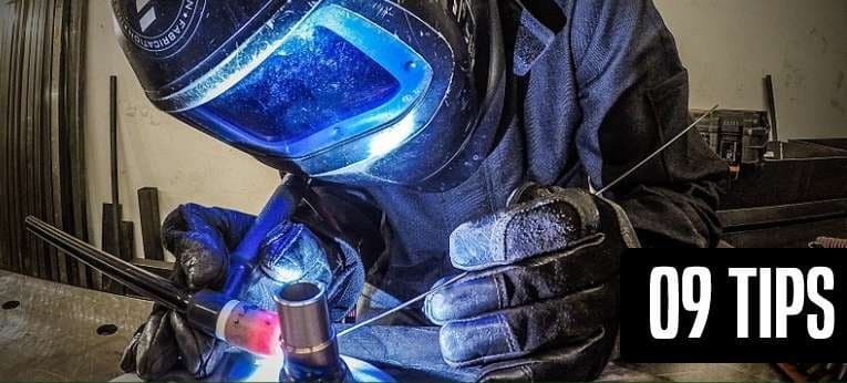 Gloved hands holding a TIG torch to a steel joint while feeding filler rod, seen over the welder's shoulder