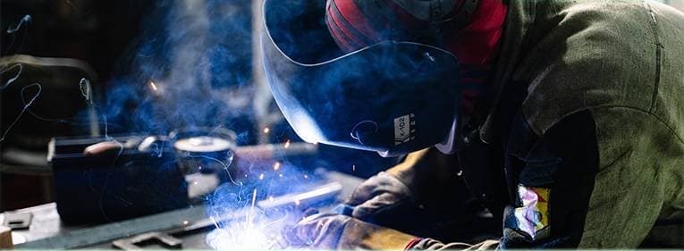 A welder in an olive jacket and red helmet leaning close to the arc, smoke rising from the bench