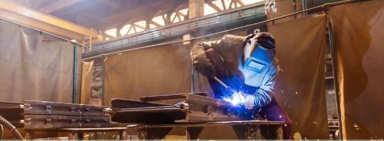 A welder in a blue helmet running a bead on steel sections at a bench, welding curtains hung behind