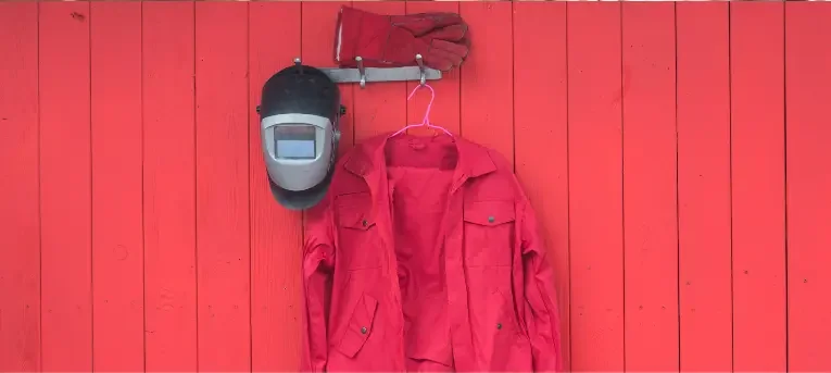 A red welding jacket, helmet and gloves hanging on a red plank wall