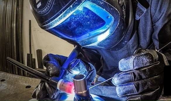 Gloved hands holding a TIG torch to a steel joint while feeding filler rod