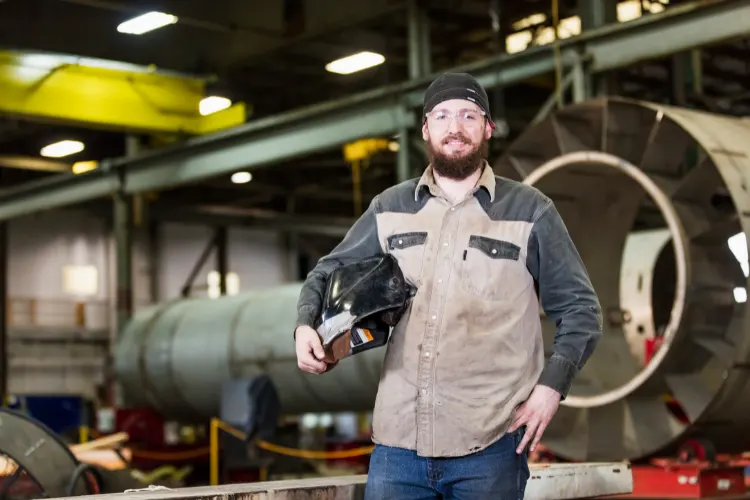 A welder in a tan flame-resistant work shirt carrying his hood through a fabrication shop