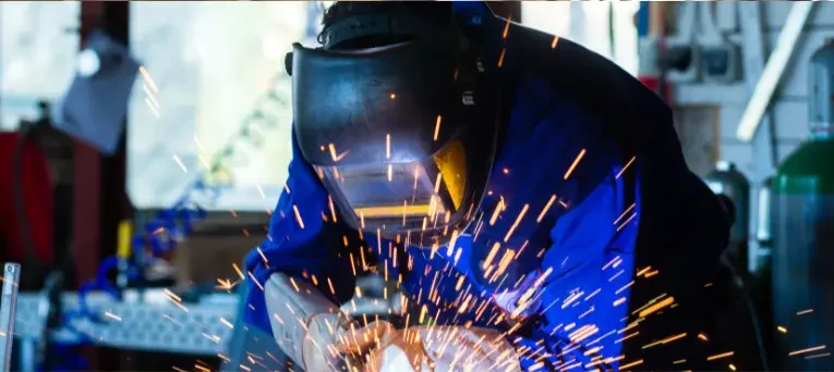 A welder in protective clothing working at a bench with sparks flying from the arc