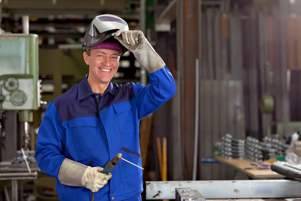 A welder in blue flame-resistant coveralls and gloves lifting his visor in a fabrication shop