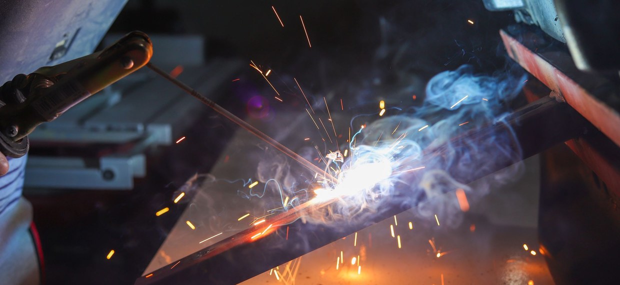 A stick electrode clamped in an electrode holder burning into steel, spatter and smoke rising, the welder's handheld shield at the edge of the frame