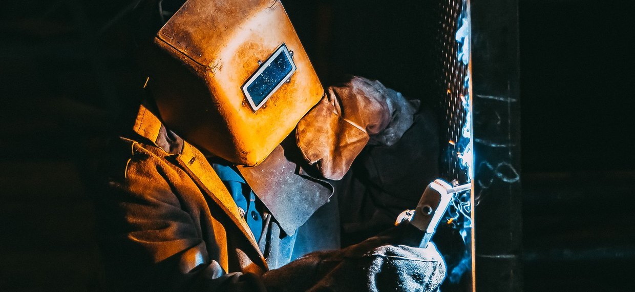 A welder in a leather jacket and shoulder cape, helmet down, striking an arc against a steel column